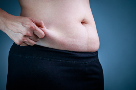 Close up female checking fat on abdomen. Woman's body with flabby stomach holding the excessive belly fat in her hand. Obesity and Overweight Concept.の写真素材