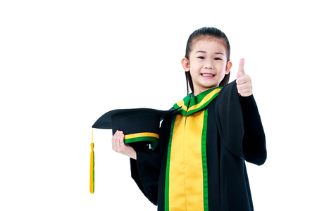 Kindergarten graduation. Happy asian child in graduation gown holding cap and showing thumb up sign at studio. Cute girl smiling happily. Isolated on white background.の写真素材