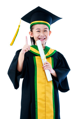 Kindergarten graduation. Asian child in graduation gown holding diploma certificate and showing forefinger up sign at studio. Cute girl gets good idea and smiling happily. Isolated on white backgroundの写真素材