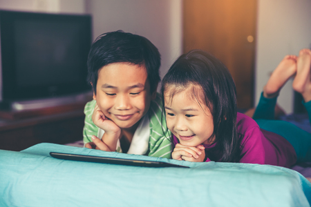 Asian children playing game on digital tablet together. Chinese boy and his pretty younger sister smiling and lying prone on bed with bright sunlight. Loving of sibling concept. Vintage film filter effect.の写真素材