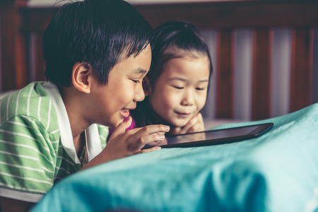 Chinese boy and his pretty younger sister smiling and lying prone on bed. Asian children playing game on digital tablet together. Conceptual about loving of sibling. Vintage film filter effect.の写真素材