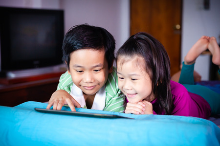 Asian children watching digital tablet for learning together. Chinese boy and his younger sister smiling and lying prone on bed. Conceptual about using E-learning technologies for educational at home.の写真素材