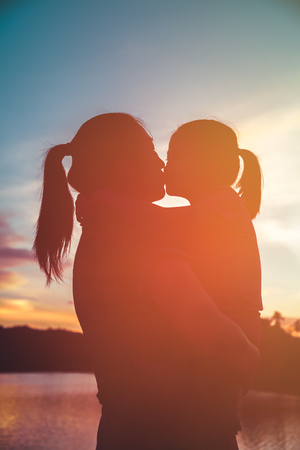 Silhouette of mother carrying and kissing her daughter. Happy family enjoying view in the morning with sunlight, cozy mood. Colorful pastel sky sunrise, serenity background. Travel on vacation.の写真素材