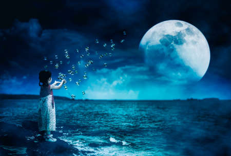 Back view of asian girl playing with soap bubbles on stone at seaside. Landscape of night sky with super moon behind partial cloud. Beautiful serenity background. The moon taken with my own camera.の写真素材