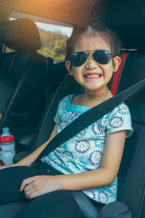 Cute asian girl in sunglasses smiling with perfect smile while sitting in the car. Tourist child relaxing, outdoors at the daytime with bright sunlight on summer day. Travel on vacation concept.の写真素材