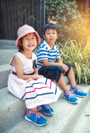 Asian children ralaxing and sitting on the stair steps. Brother and sister smiling happily, outdoor with sunlight on summer day. Happy family spending time together, loving and bonding of siblingの写真素材