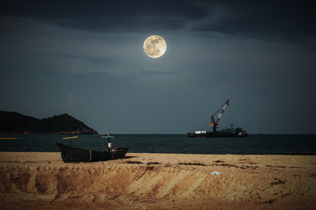 Beautiful night sky with bright full moon above cargo ship anchored in the sea. Traditional fishing boat parked on tropical beach. Outdoor at nighttime. The moon taken with my camera.の写真素材
