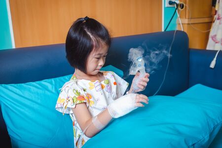 Sad asian child holds a mask vapor inhaler for treatment of asthma. Breathing through a steam nebulizer.Illness girl admitted in hospital while saline intravenous (IV) on hand. の写真素材