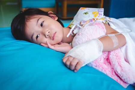 Illness asian child admitted in hospital while saline intravenous (IV) on hand. Unhappy girl depress and hopeless with absent minded looking away, kid emotion. Health care stories. の写真素材
