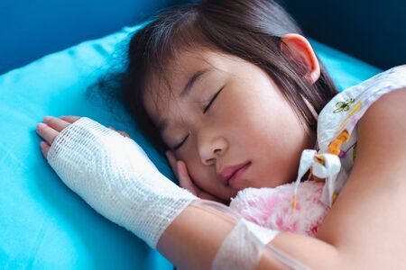 Closeup of illness asian child admitted in hospital with saline intravenous (IV) on hand. Girl sleeping at comfortable equipped hospital room. Health care stories.の写真素材