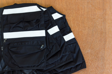 Close up of many black vest with reflective strips are stacked on white wooden floor.
