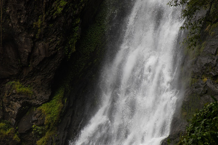 Close-stream waterfall which flows swiftly through severe cliff rocks with ferns, moss covered...