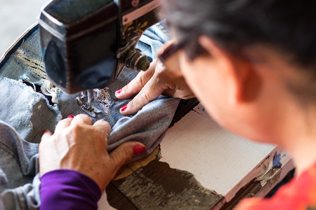Rear view of an elderly woman doing repairs on a pair of jeans on an old sewing machine.の写真素材