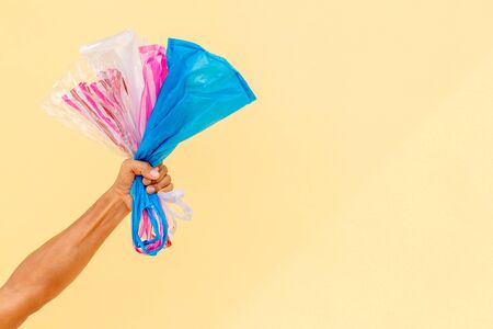 Many colorful plastic bags crumpled in the hands of a man with a yellow concrete wall in the background.の写真素材