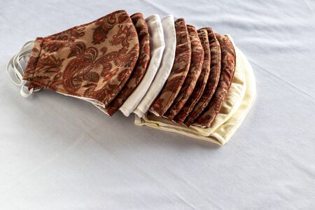 Close up, many mouth masks and Thai patterned masks are stacked on a white piece of fabric which is...