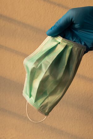 Close up, hand in blue gloves, holding a green mask with a concrete wall and the sun shining through...