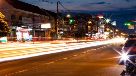 The abstract of the car headlights stretches over an asphalt road near a row house at night in the Thai province of town.の写真素材