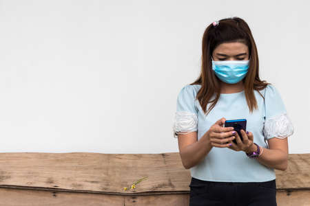 A close-up portrait of a Thai woman wearing a blue face mask playing mobile phone near an old wooden...