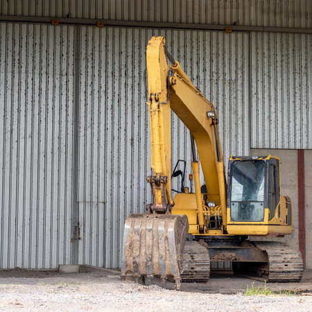 A yellow backhoe is parked close to the galvanized wall of a factory in Thailand.