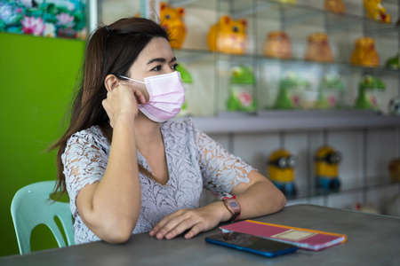 Close-up of a portrait of a Thai woman wearing a pink mask sitting at a table weary of the coronavirus covid-19 at a table inside her shop.の写真素材