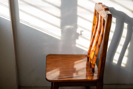 A close-up view of a wooden chair standing alone in a corner of the room where sunlight shines through the shuttered windows onto the white concrete walls.の写真素材