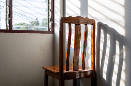A close-up view behind the back of a wooden chair, standing alone in a corner of the room, where sunlight shines through the shuttered windows onto the white concrete walls.の写真素材