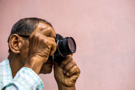 Close-up of both hands of an elderly Thai man holding an old film camera covering his face and staring to take a picture with a pink concrete wall in the background.の写真素材