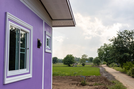 A view through the side of a contemporary purple house towards green rice fields and Thai countryside roads with cloudy afternoons.の写真素材