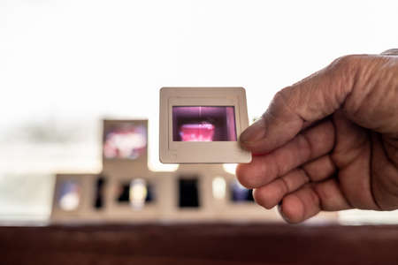 A close-up view of the 35mm slide film in middle-aged fingers with a group of rear slides backlit on a framed glass louvered window to reminisce about the past.の写真素材