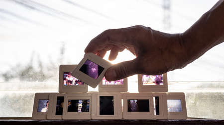 A close-up view of the 35mm slide film in middle-aged fingers with a group of rear slides backlit on a framed glass louvered window to reminisce about the past.の写真素材