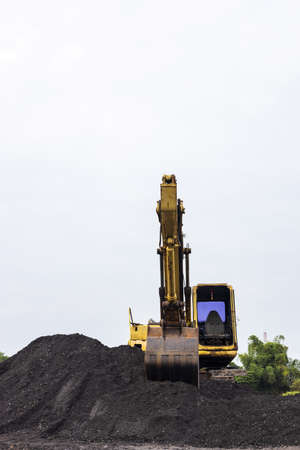Vertical low view in front of an old yellow backhoe parked on a mound of black soil with trees far...