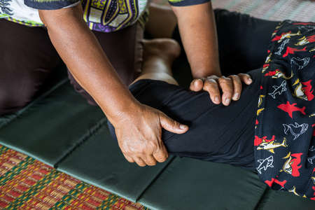 Close-up view of both hands of an elderly Thai female masseuse squeezing and bending the legs of another elderly woman lying on a mat to relax and relieve the pain.の写真素材