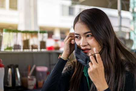 Take a close-up portrait of a beautiful Thai woman about to take off a black mask worn to prevent...