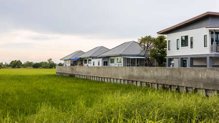 View of a modern residential housing estate with concrete fence walls that stretch over a green rice field in the Thai countryside during cloudy day.の写真素材
