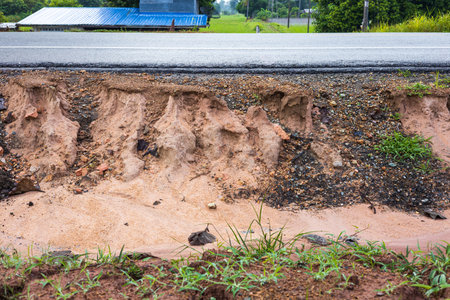 A side view of the sandstone soil beneath an asphalt road eroded by rain and wild water, with residential houses and rice fields as a backdrop in rural Thailand.の写真素材