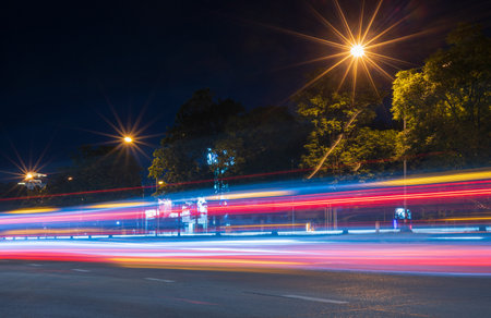 A view from the side of a road where car lights pass by from a long exposure are abstract red-white stripes near trees and light from Thai suburban lamps.の写真素材