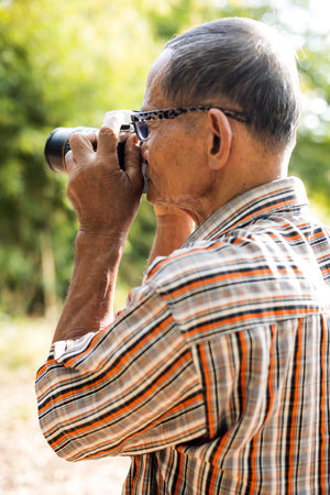 Side shot of an elderly Thai man wearing glasses holding an old DSLR camera and looking at something to press the shutter in rural Thailand.の写真素材