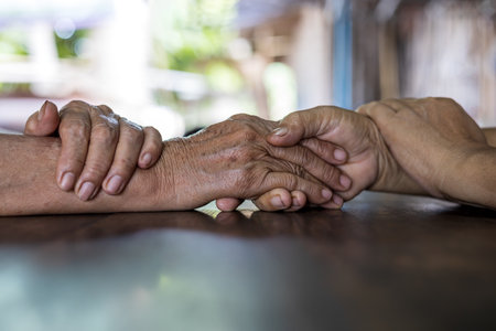 Low angle close-up shot of two wrinkled hands of two elderly Thai women holding each other in consolation on an old wooden table with sunlight reflections outside the house in the background.の写真素材