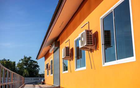 Close-up low-angle view of an orange building with sliding glass windows and an air conditioner compressor mounted near a steel fence on a concrete floor and a blue sky in the afternoon as a backdrop.の写真素材