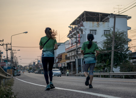 A low-angle view of the back of runners running on paved roads competing over bridges and intersections with residential buildings in early morning Thailand.の写真素材