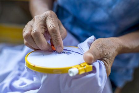 Close-up view. Hand of an elderly Thai woman using a slitting needle to pull the old blue thread from which the letters are embroidered out of the white cloth stretched taut with a plastic hoop.の写真素材