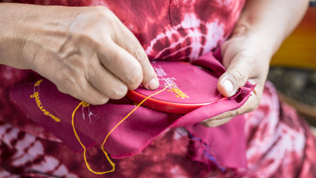Close-up view of hands of an elderly Thai woman wearing a red blouse using a needle and yellow...