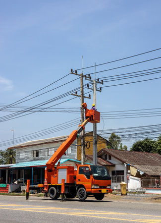 An orange crane truck belonging to the power unit lifts a lift to send people to repair power lines on a power pole during daylight hours near a house in rural Thailand.の写真素材