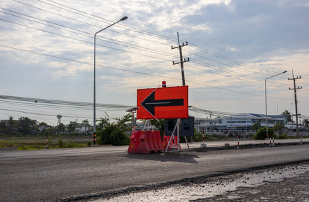 A view of the surface of a new black asphalt road with signs and orange barricades placed in the middle of a road being renovated near a power pole in rural Thailand during the day.の写真素材