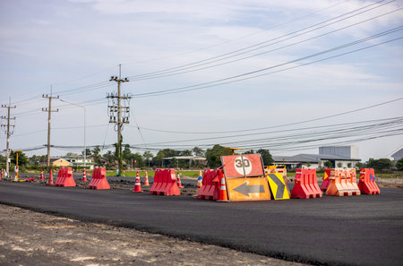 A view of the surface of a new black asphalt road with signs and orange barricades placed in the middle of a road being renovated near a power pole in rural Thailand during the day.の写真素材