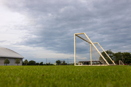 Low angle view of an old white metal soccer goal installed on a grass field near a factory building with overcast clouds and rising sun in the background in a rural Thai countryside.の写真素材