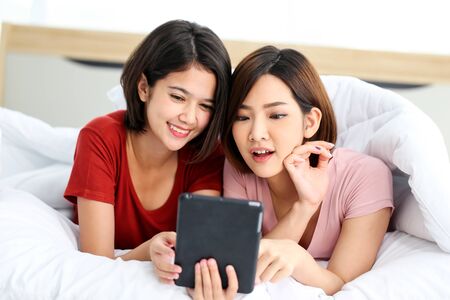Happy young couple friend beautiful asia women lying on the bed in a bedroom and using digital tablet.の写真素材