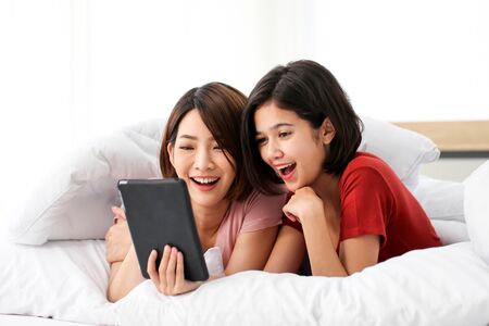 Happy young couple friend beautiful asia women lying on the bed in a bedroom and using digital tablet.の写真素材