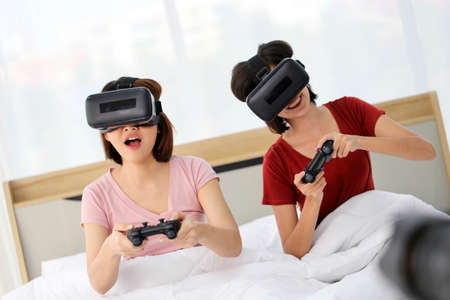Happy young couple friend beautiful asia women playing video games virtual reality glasses on the bed in a bedroomの写真素材