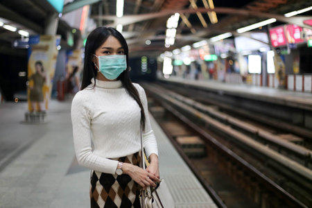 Beautiful asia woman passenger wearing medical hygiene protective face mask infection from coronavirus covid-19 standing in lines waiting sky train on platform at night.の写真素材